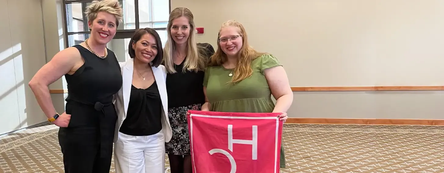 Four students posing with a pink HerCampus club flag.